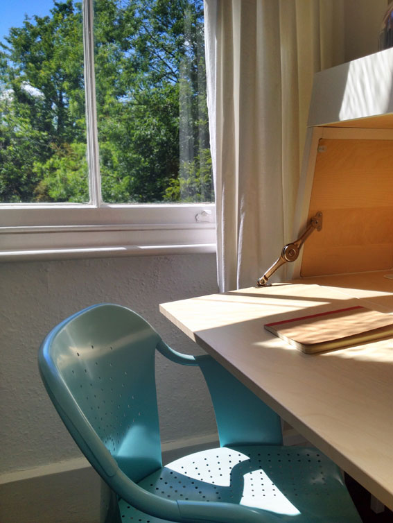 Opened desk with light blue chair and window overlooking mature trees and blue sky